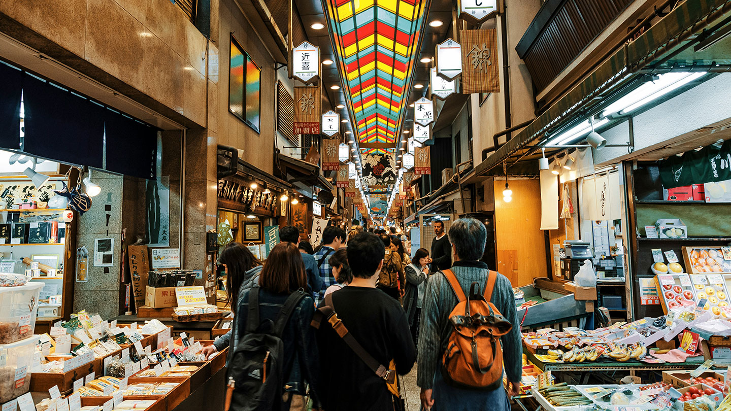 カラフルなアーケード屋根の下、さまざまな店舗が並ぶ商店街を多くの人が歩いている