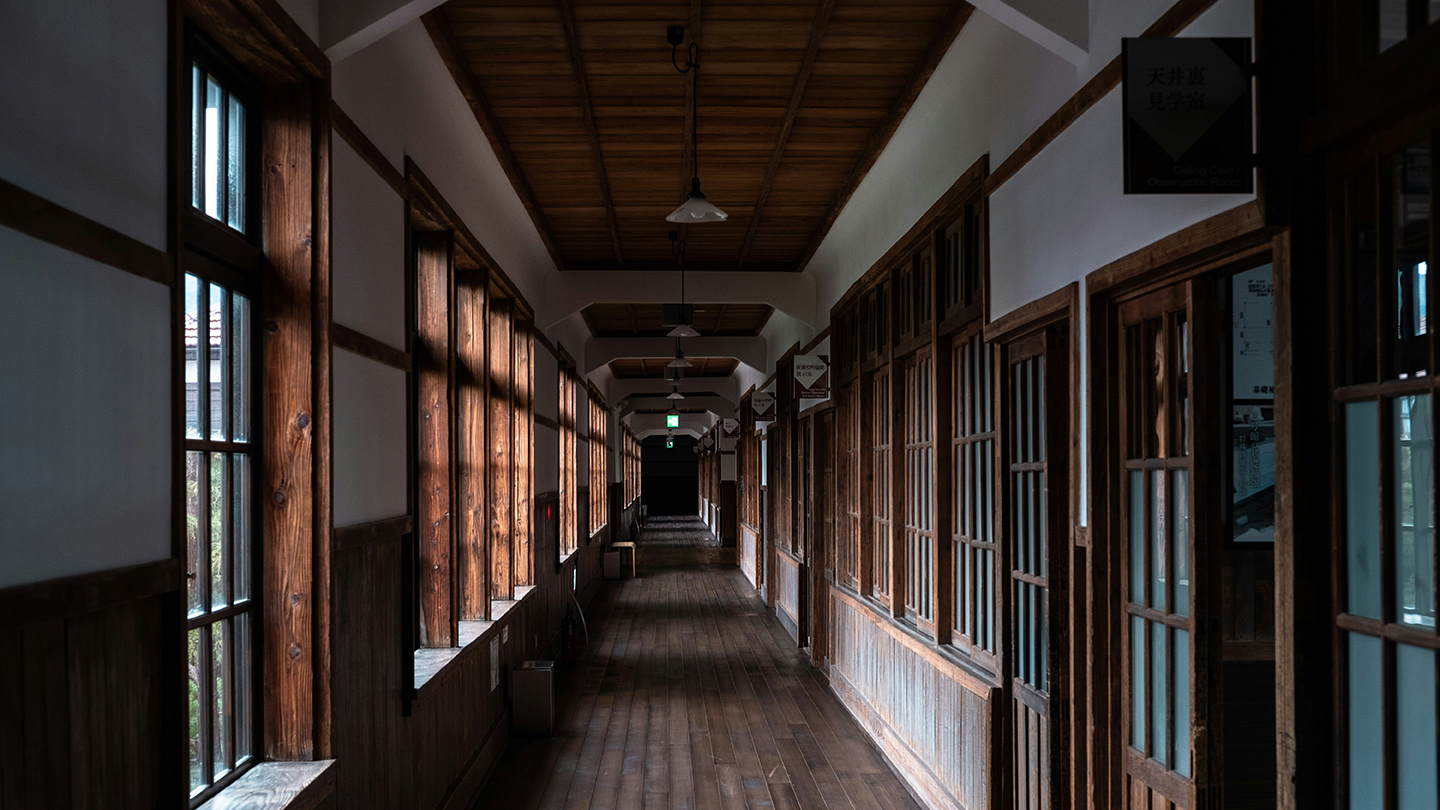 木造校舎の長い廊下に、木枠の窓や教室の扉が並び、奥へ向かってまっすぐ続いている