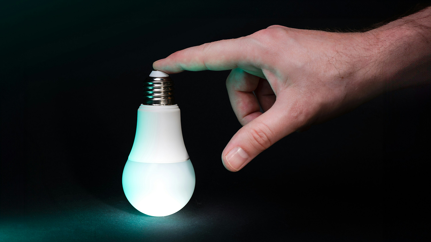 黒い背景の前で、点灯した電球型のライトに指先で触れようとしている手