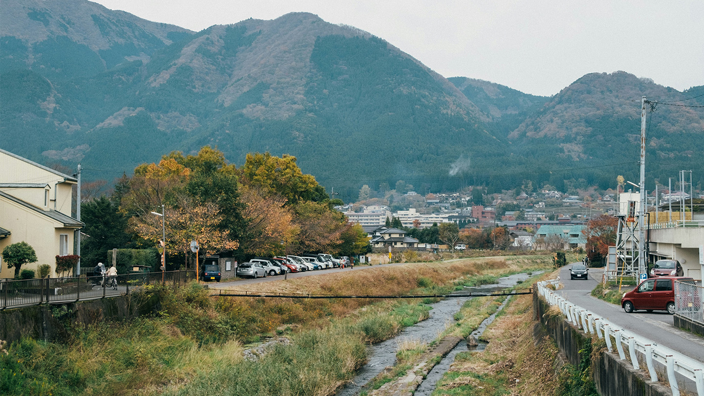 山あいの集落を流れる川沿いに、住宅や道路、駐車場が広がっている。背後にはなだらかな山並みが見える