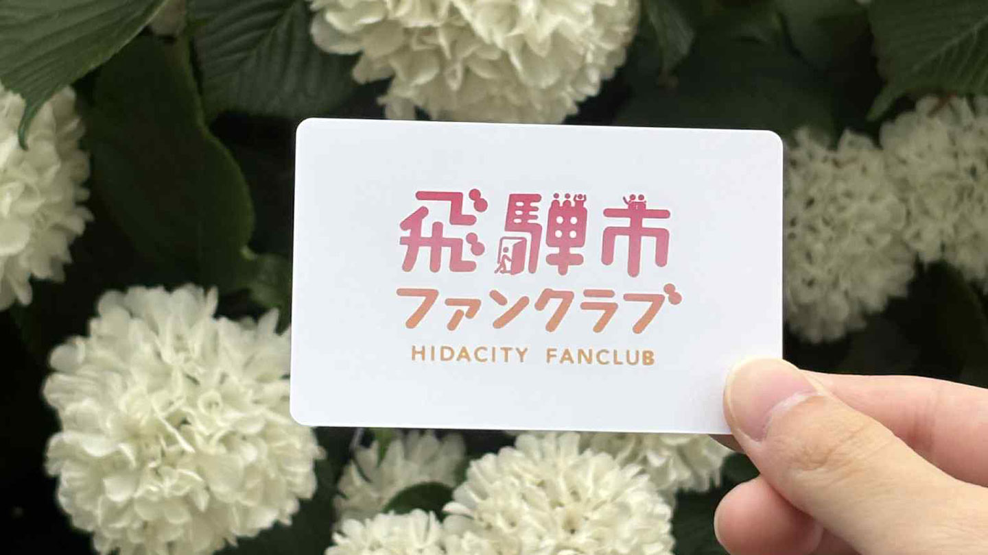白いアジサイの花を背景に、「飛騨市ファンクラブ」と記されたカードを手に持つ様子