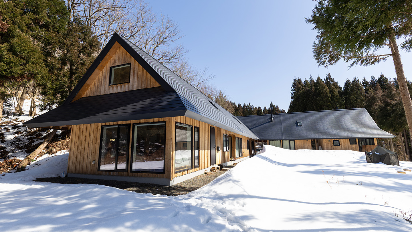 雪に覆われた敷地に建つ、黒い屋根と木の外壁が特徴的な平屋の建物