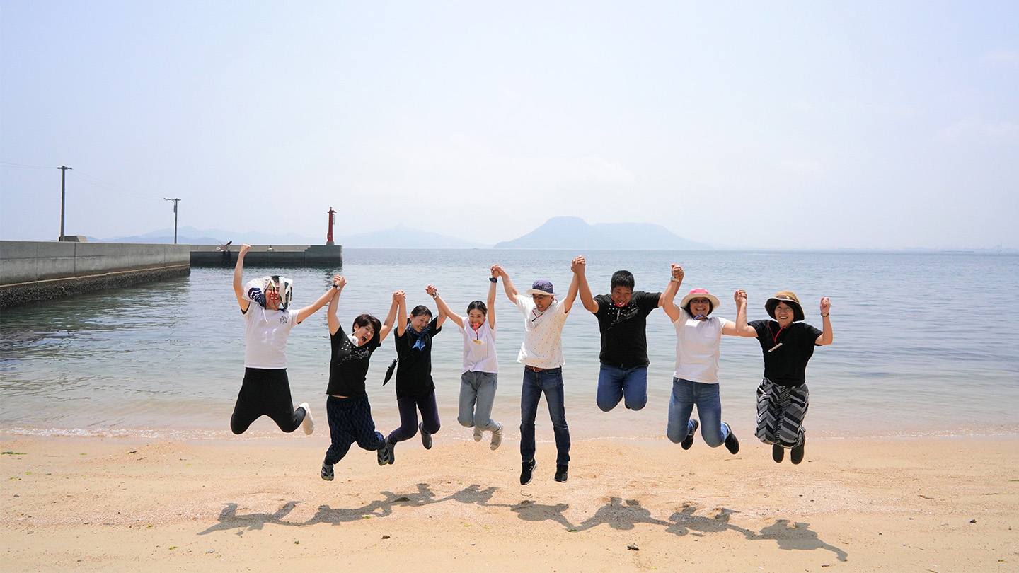 海を背景に、砂浜で複数人が手をつないでジャンプしている
