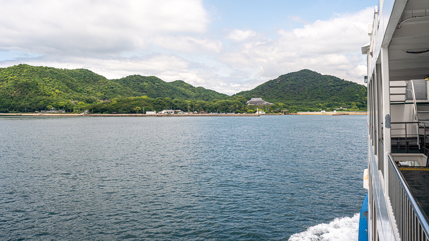 船上から海と島の山並みを望む風景。右側に船体の一部が写り、海面には白い波が立っている