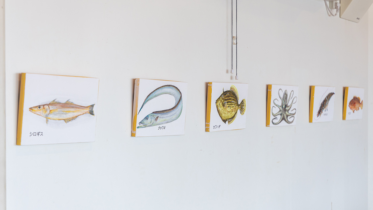 白い壁に、魚やタコなど海の生き物を描いた小さな絵が横一列に展示されている