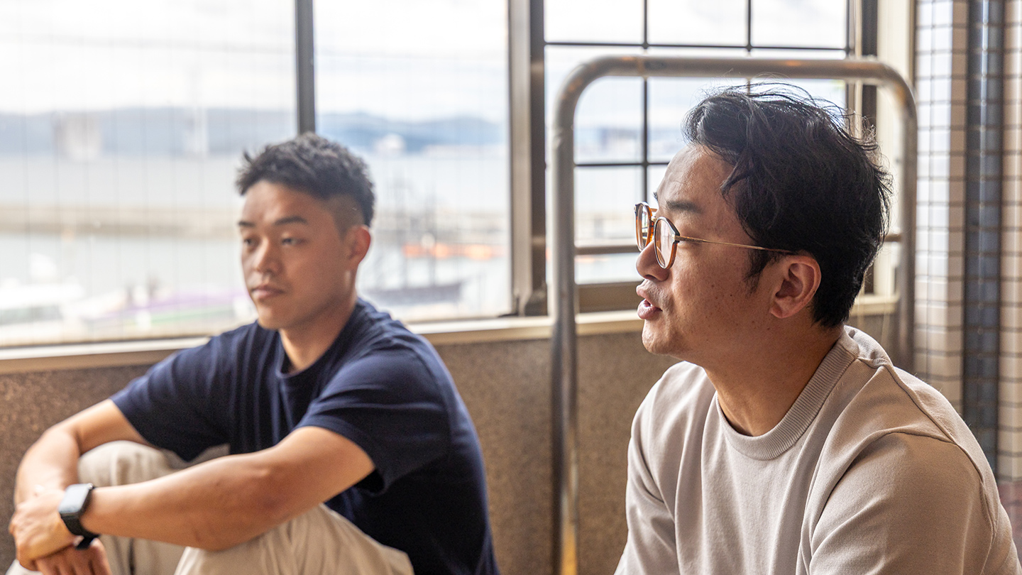 屋内で窓辺に座って話をしている大石一仁さんと湯川致光さん