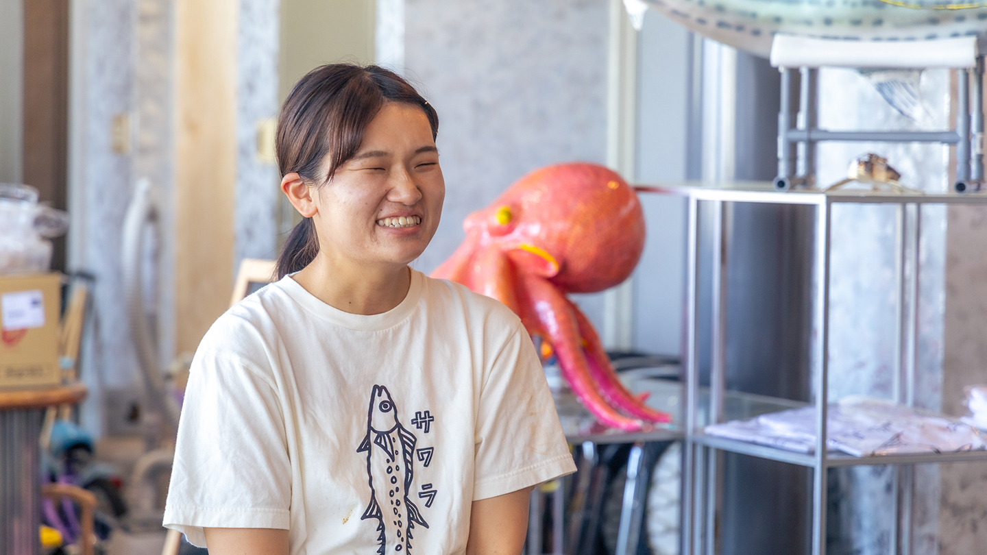 屋内で大石佑紀さんが笑顔で話している。背景には魚やタコをかたどった展示物