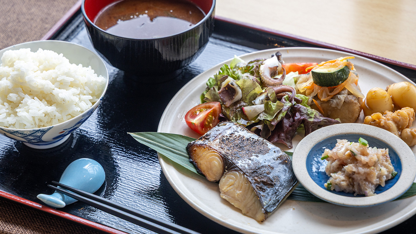 焼き魚やサラダ、小鉢、ご飯、味噌汁が黒い盆に並んだ定食