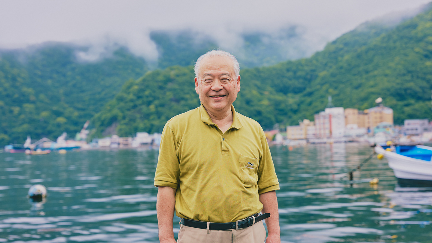 海と山、漁港を背景に、笑顔で中島さんが立っている