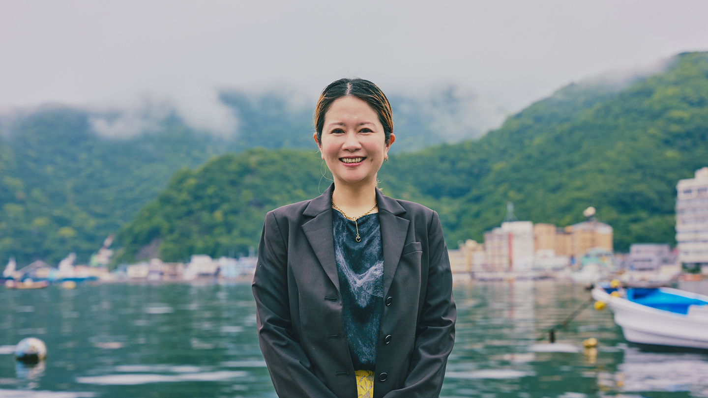 海と山、漁港を背景に、青山さんが笑顔で立っている