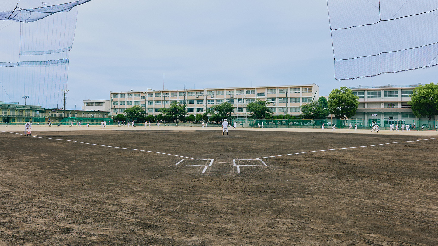 グラウンドで白い練習着姿の野球部員たちが練習し、その様子を藤井監督が見守っている