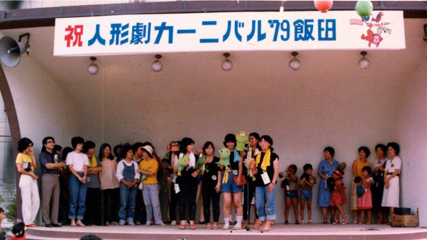 「祝人形劇カーニバル’79飯田」と書かれた看板。その下のステージに、人形を持って立つ人たち