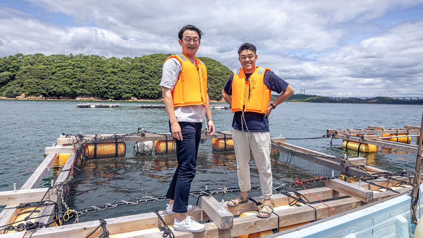 海上の生簀の上で、オレンジ色のライフジャケットを着た湯川致光さんと大石一仁さんが並んで立っている。背景には海と緑の島、曇り空が広がる