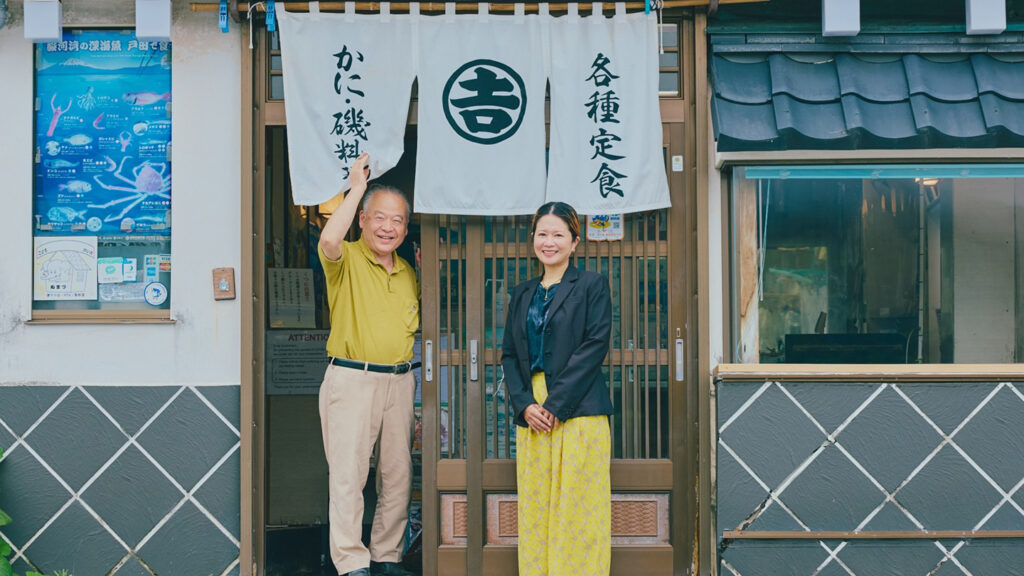 丸吉食堂の暖簾をあげる中島寿之さんと横に立っている青山沙織さん