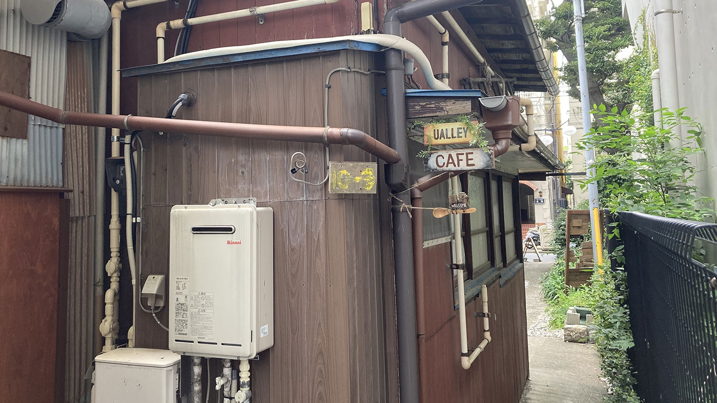 古い木造建物の外壁に、配管や給湯器が取り付けられている。右側には建物の間の細い路地が続いている