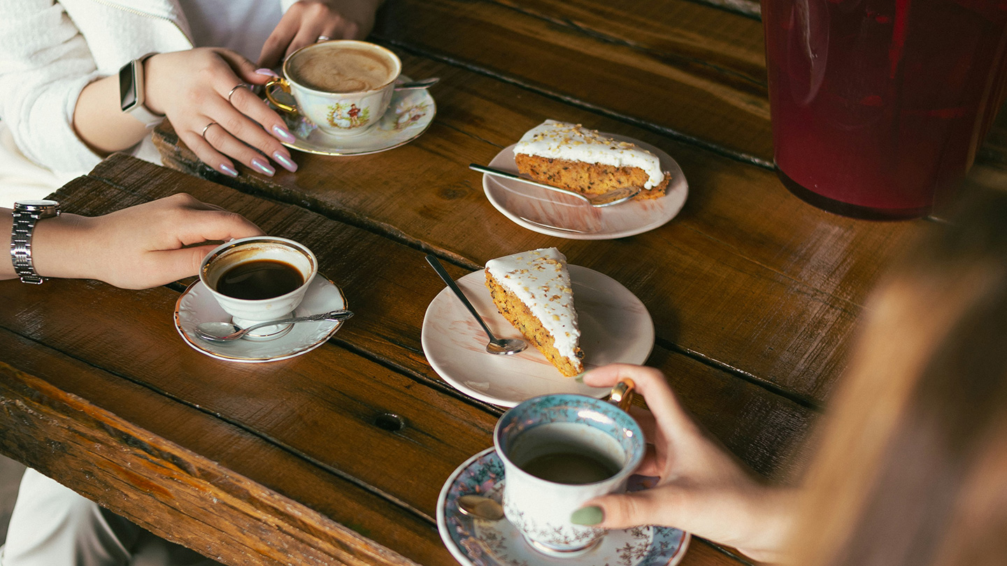 木製のテーブルに並ぶコーヒーとケーキを囲んで、数人がカフェで過ごしている