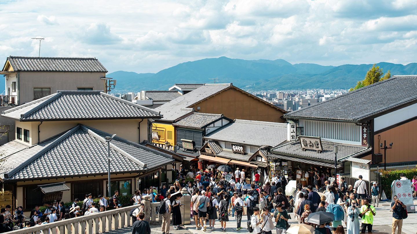 山並みを望む観光地の通りに、瓦屋根の建物が並び、多くの観光客が行き交っている