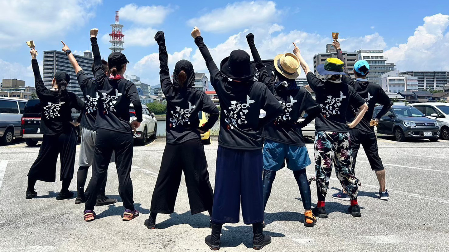 青空の下、後ろを向いた8名の踊り子が左手を空に向かって突き上げている