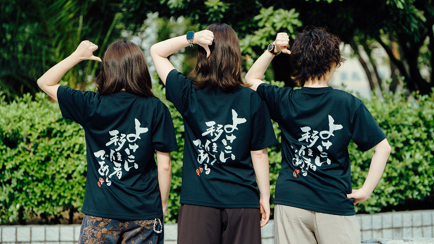 山本さん、芳村さん、足立さんが並んで後ろを向き、黒いTシャツの背中に白い文字で書かれた「よさこい移住者である」という言葉を親指で指している
