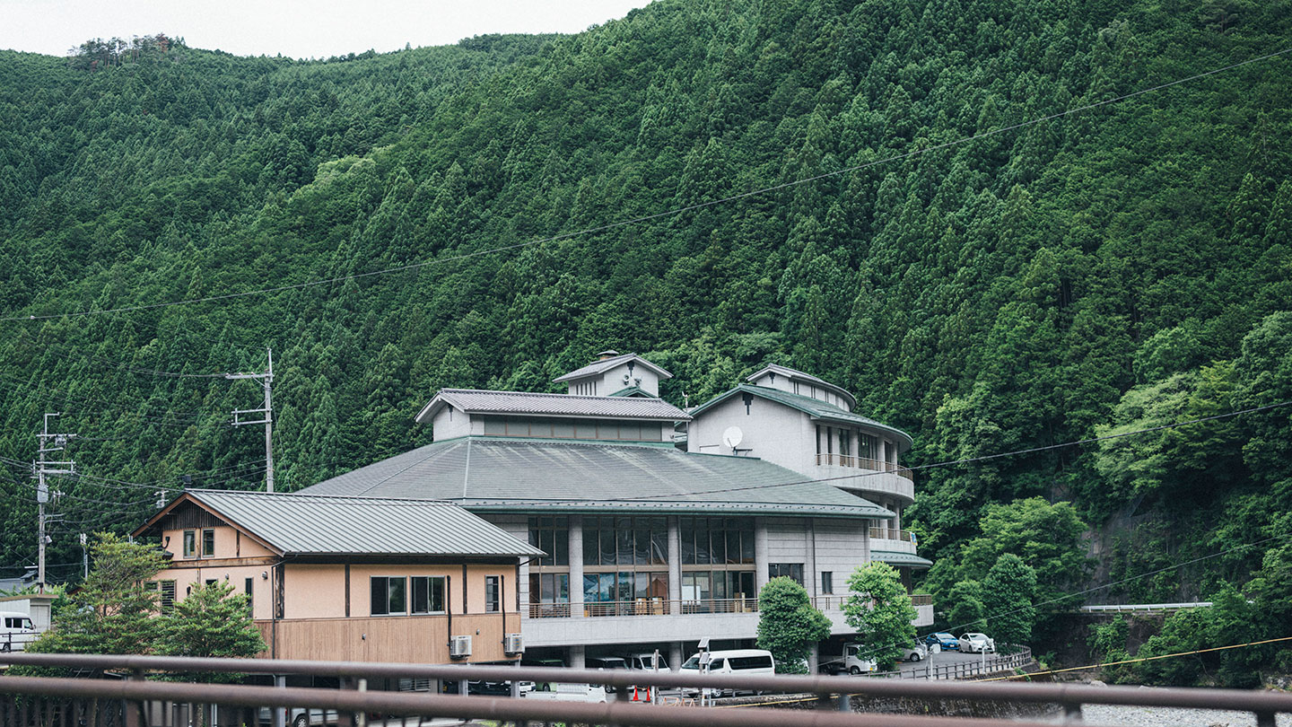 大きな建物の背後に、緑の木々に覆われた山が広がっている