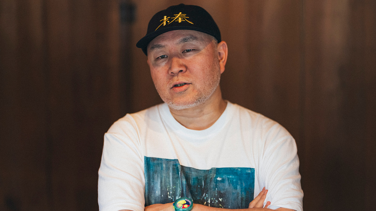 黒いキャップをかぶった坂本大祐さんが、室内で話している