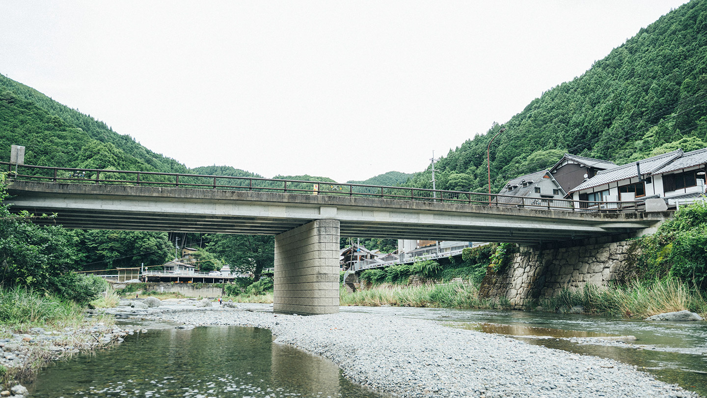 山あいを流れる川に橋がかかり、周囲に緑の山々と集落が広がっている