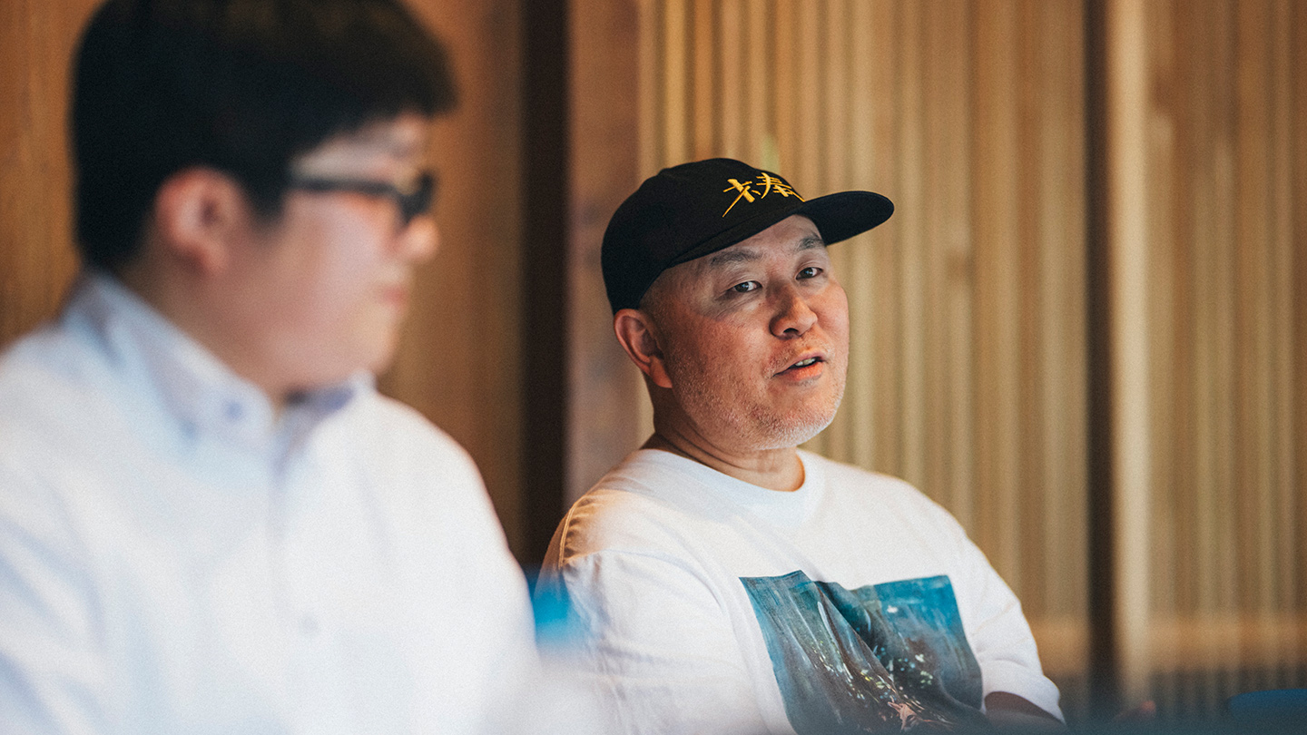 黒いキャップをかぶった坂本大祐さんが、室内で話している
