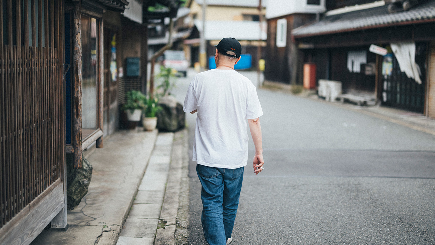 古い建物が並ぶ通りを、白いTシャツ姿の坂本さんが歩く後ろ姿