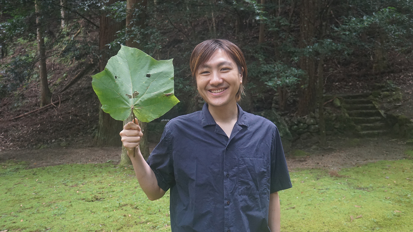 青木真兵さんが、大きな葉を手に笑顔を見せている