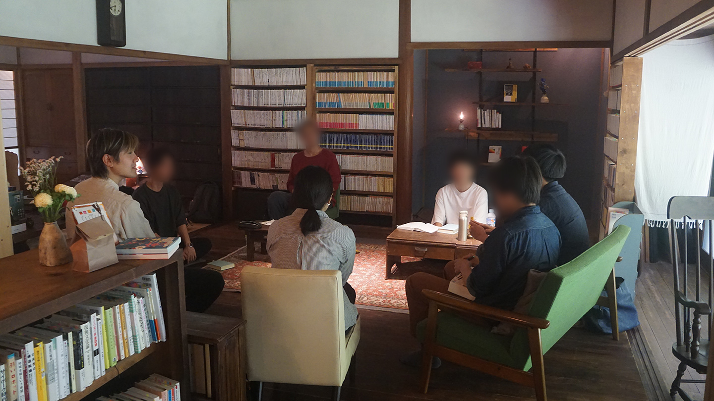 本棚のある室内で、複数の人物が輪になって座り、読書会をしている
