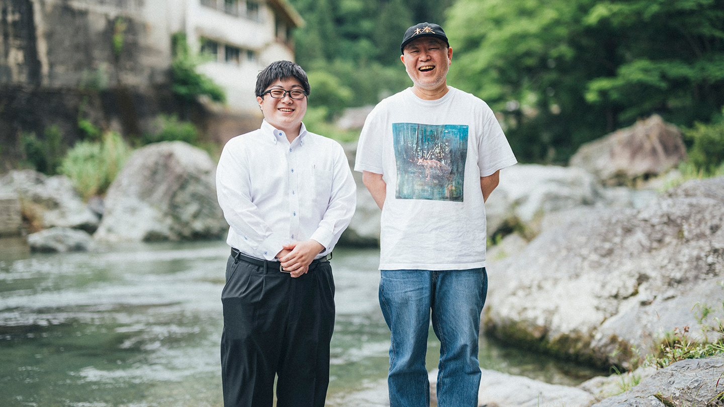 川辺に立つ北川拓哉さんと坂本大祐さんが、緑に囲まれた自然のなかで笑顔を見せている