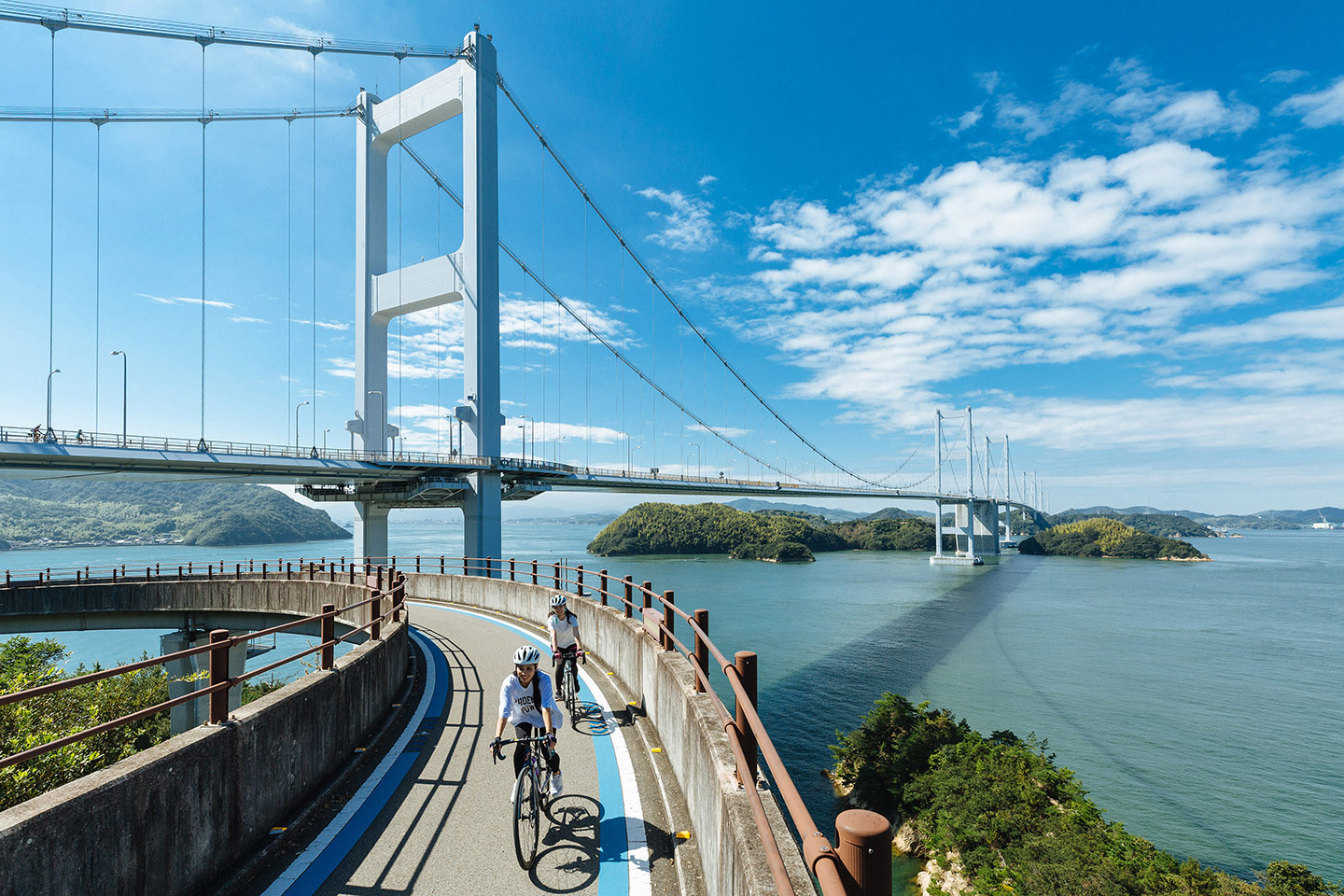青い空と海が広がる瀬戸内海で、島々を結ぶ吊り橋を背景に、海沿いのサイクリングロードを自転車で走る二人