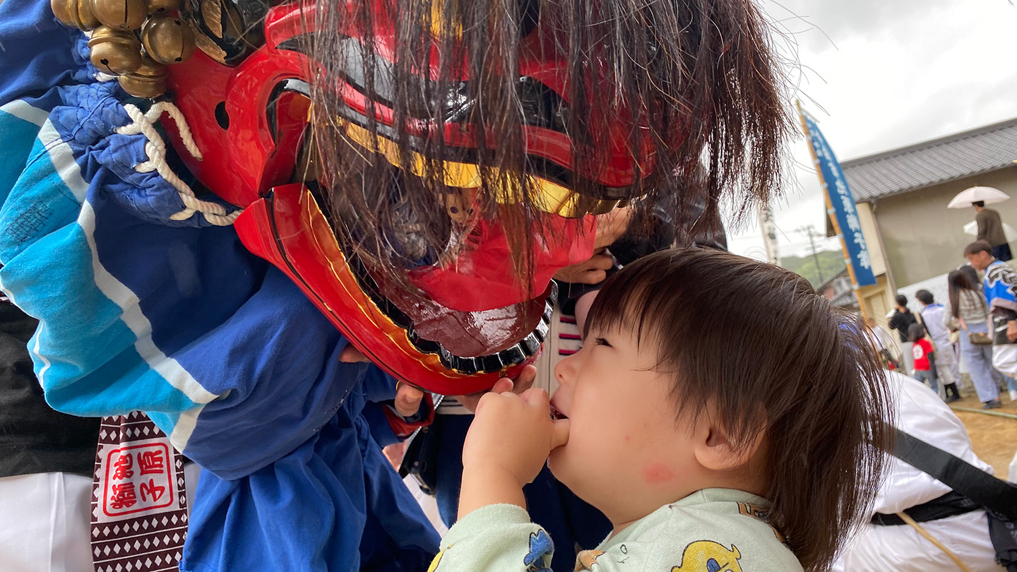 祭りの会場で、子どもの頭に獅子舞が顔を近づけている