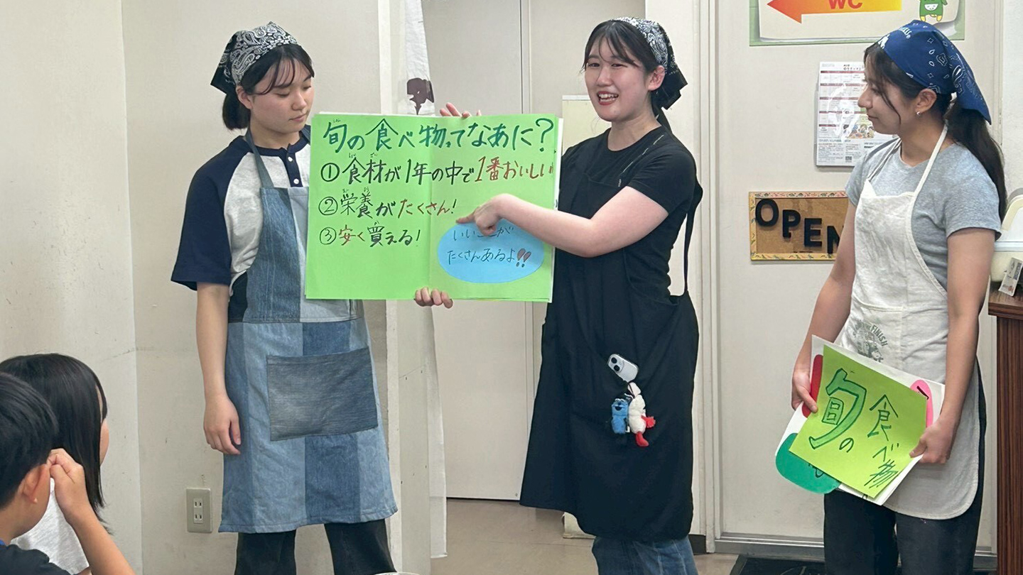 エプロンをした3人の学生が、子どもたちの前で旬の食べ物の説明をしている