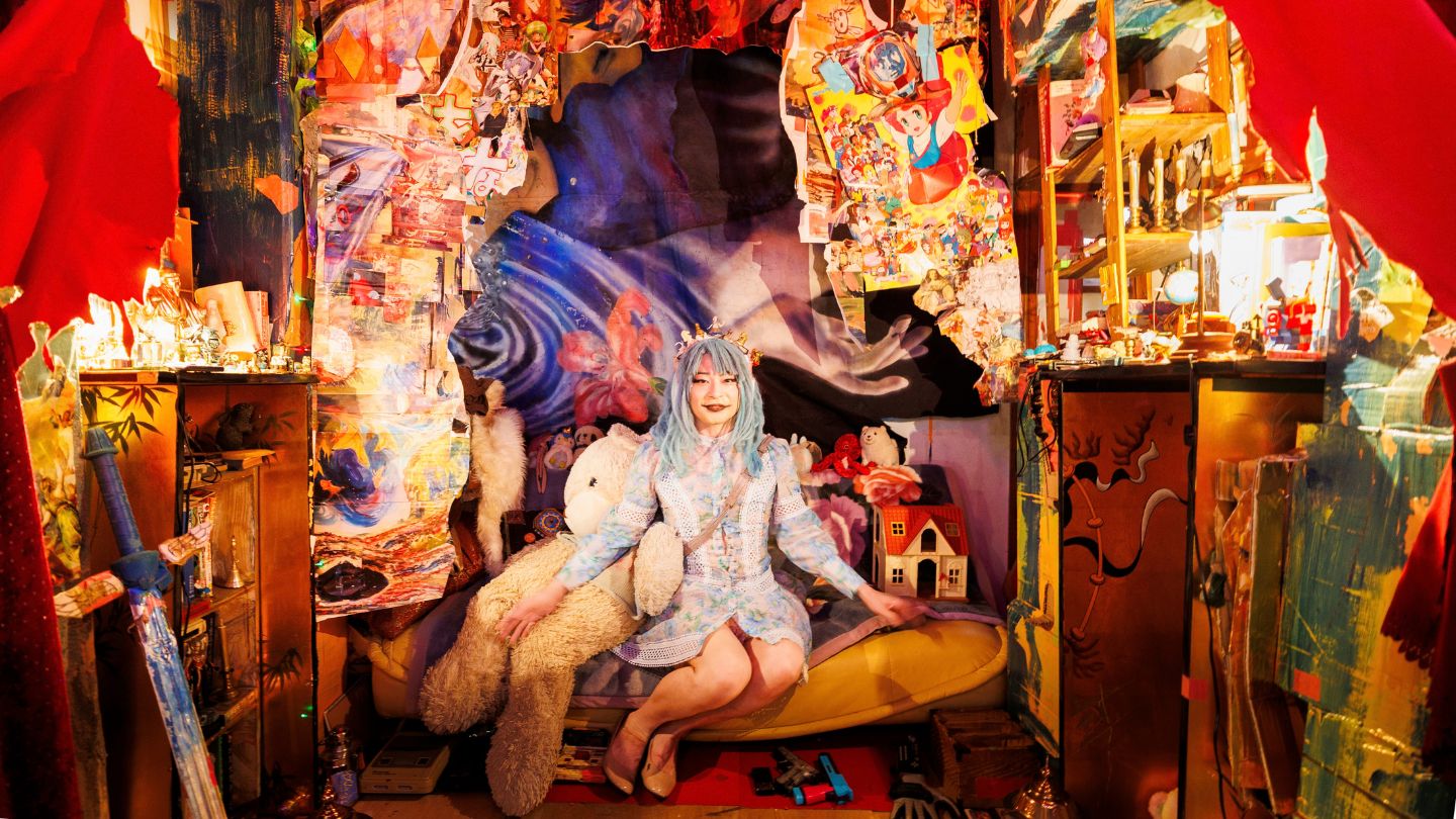 色とりどりの作品で埋め尽くされたアトリエ。メイクと衣装で着飾った東京ディスティニーランドさんが、大きなクマのぬいぐるみと並んで黄色いソファに座っている