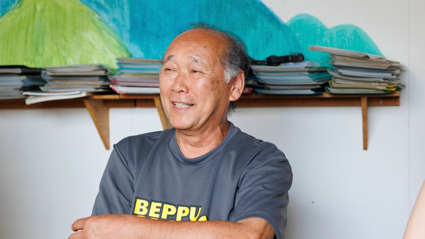 本と山の絵画を背景に、黄色いBEPPUの文字がプリントされたTシャツを着て話す山本さん