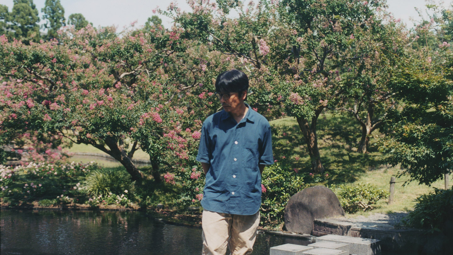 庭園の池のほとりに佇む岸田繁さん。背景にピンク色の花を咲かせたサルスベリの木々が広がる