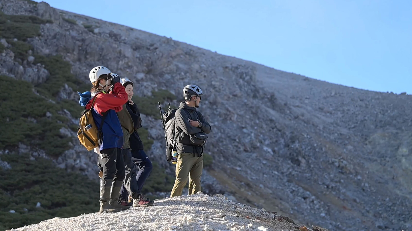 岩肌が広がる山中で、ヘルメットと登山装備を身につけた3人が周囲の景色を眺めている