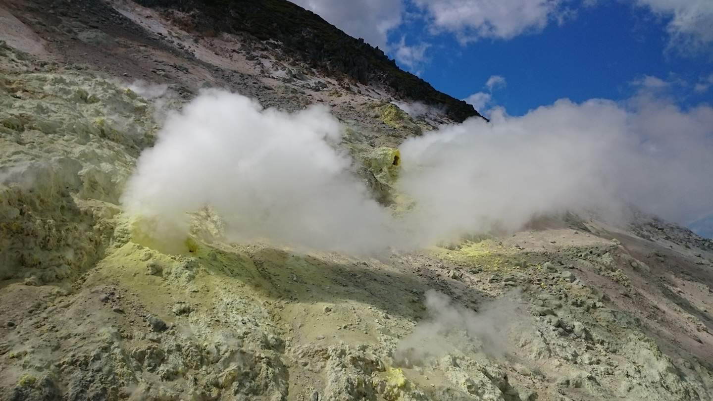 岩肌の噴気孔から白い蒸気が立ちのぼり、周囲に黄色い硫黄が広がっている