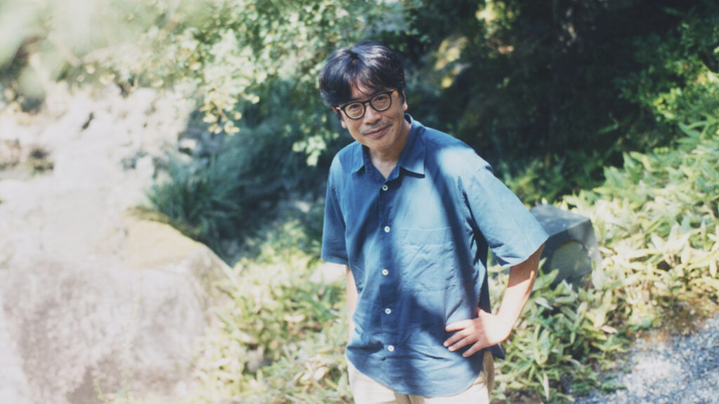 日本庭園のなかで、腰に手を当てて微笑む岸田繁さんのポートレート