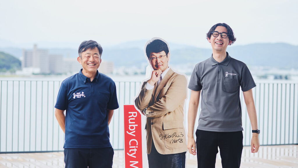 まつもとゆきひろさんのパネルの横で笑う市の担当者