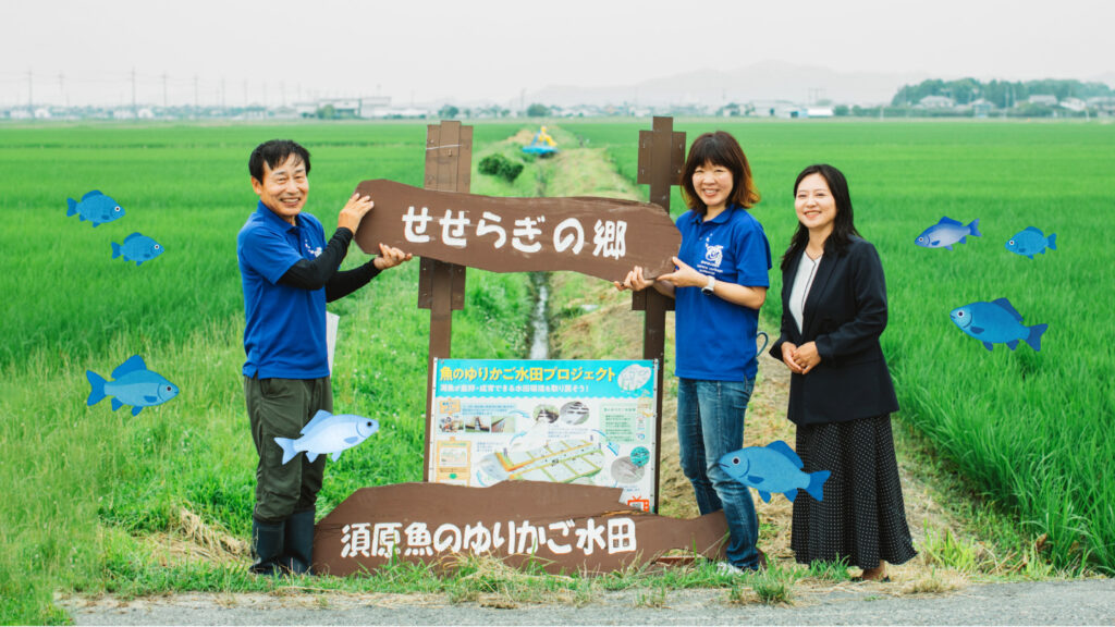 水田を背景に、辻本さんと藤岡さん、西田さんが笑顔で立っている。看板には「せせらぎの郷　須原魚のゆりかご水田」の文字