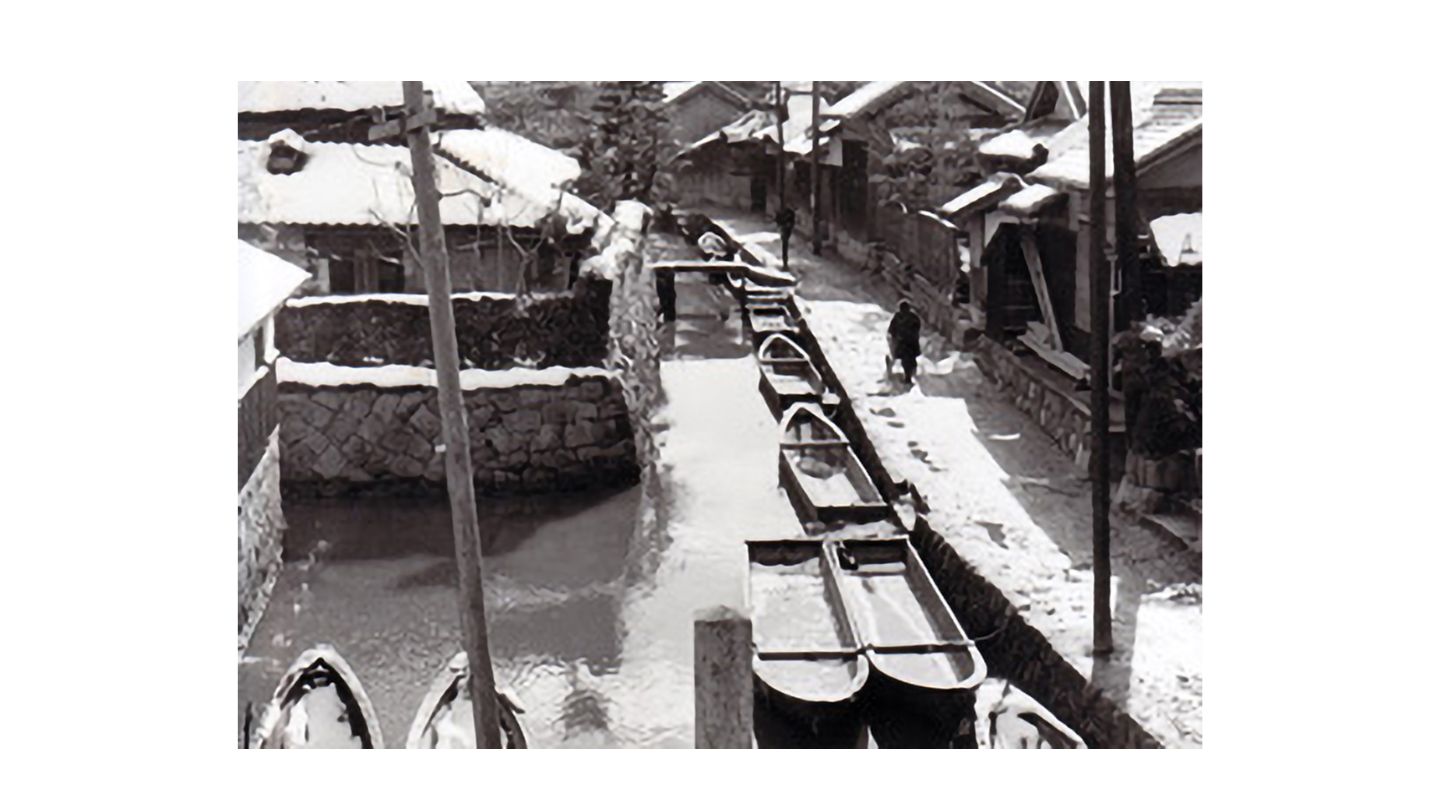住宅沿いの水路の上に田舟が並んでいるモノクロ写真