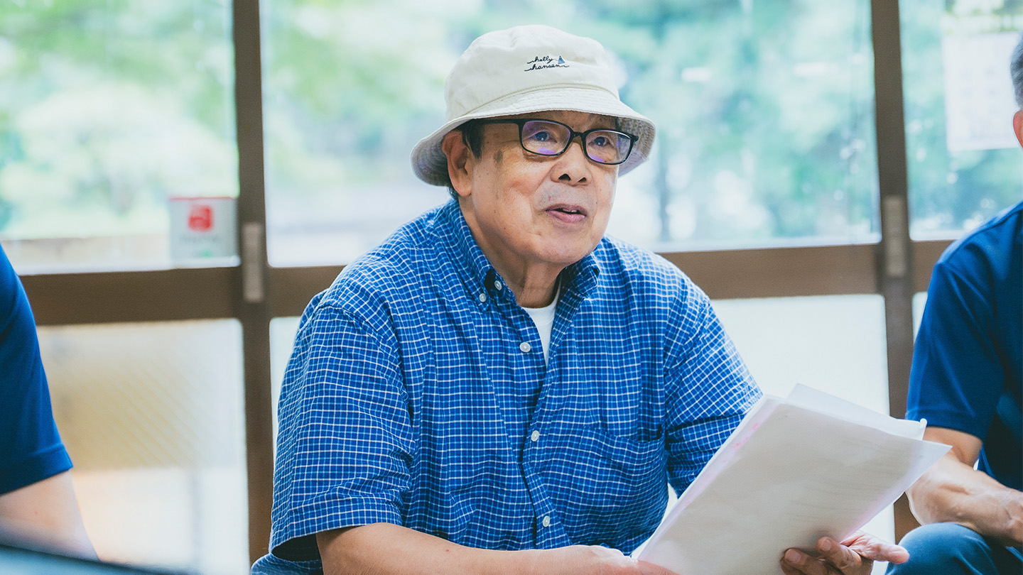 白い帽子をかぶり、青いチェック柄のシャツ姿で資料を手に話す犬井正さん