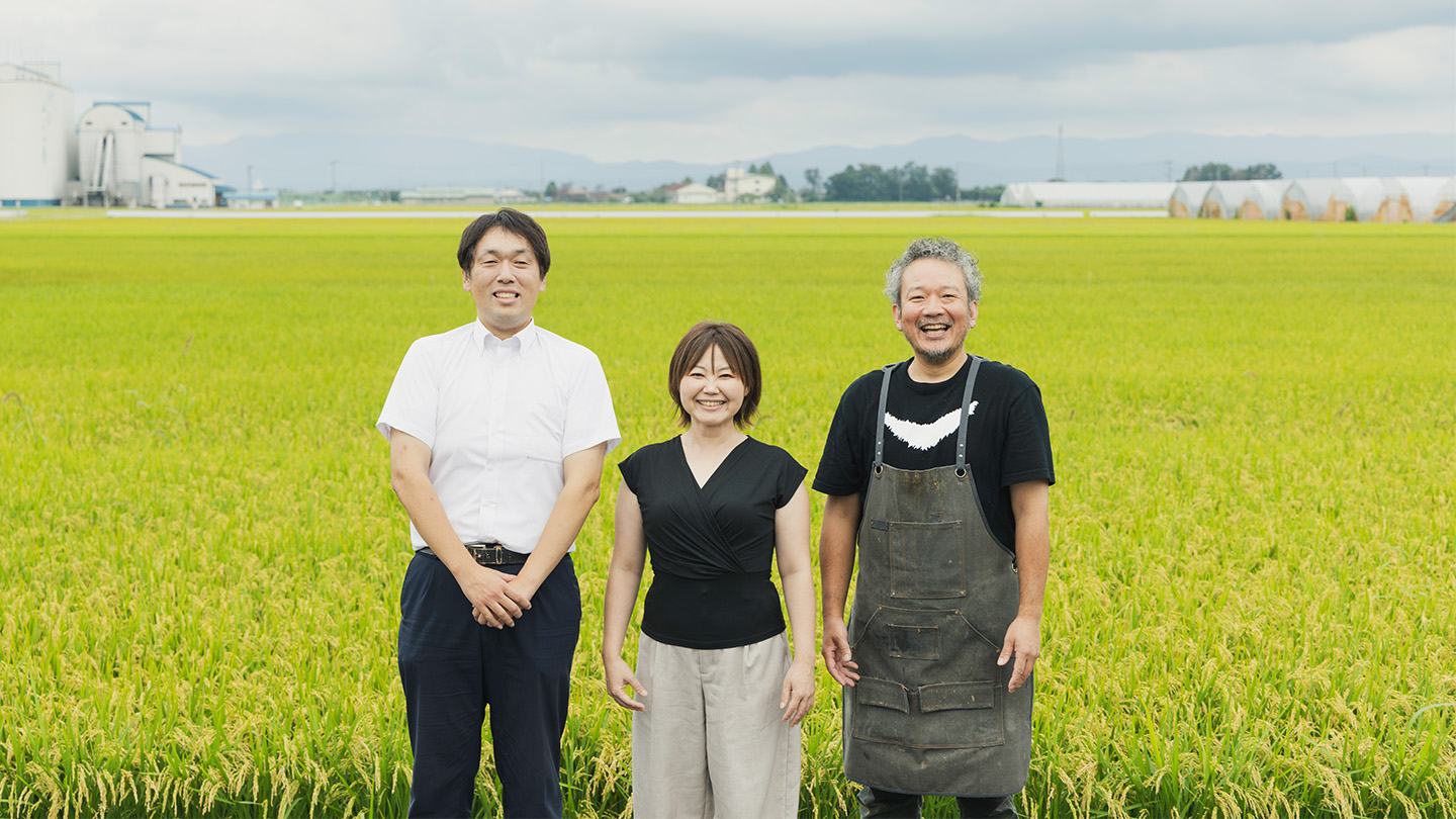 稲穂が実った田んぼを前に、小野愛美さん、齋藤光さん、シェフの古門浩二さんが笑顔を浮かべて並んで立っている